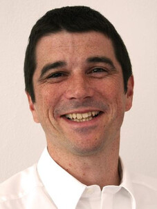 Porträtfoto von Sebastian Goennenwein freundlich lächelnd im weißen Hemd vor hellem Hintergrund.